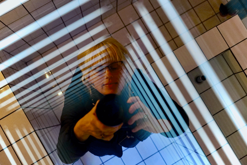 Reflection of a woman with blonde hair and glasses holding a camera, seen through striped glass panels with tiled floor background.