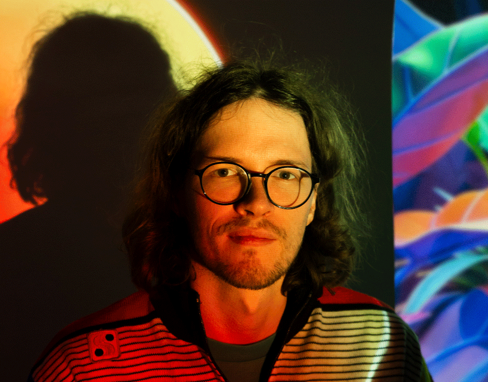 Man with shoulder-length curly hair and round glasses illuminated by colorful lights, with a shadow cast on the wall behind him.