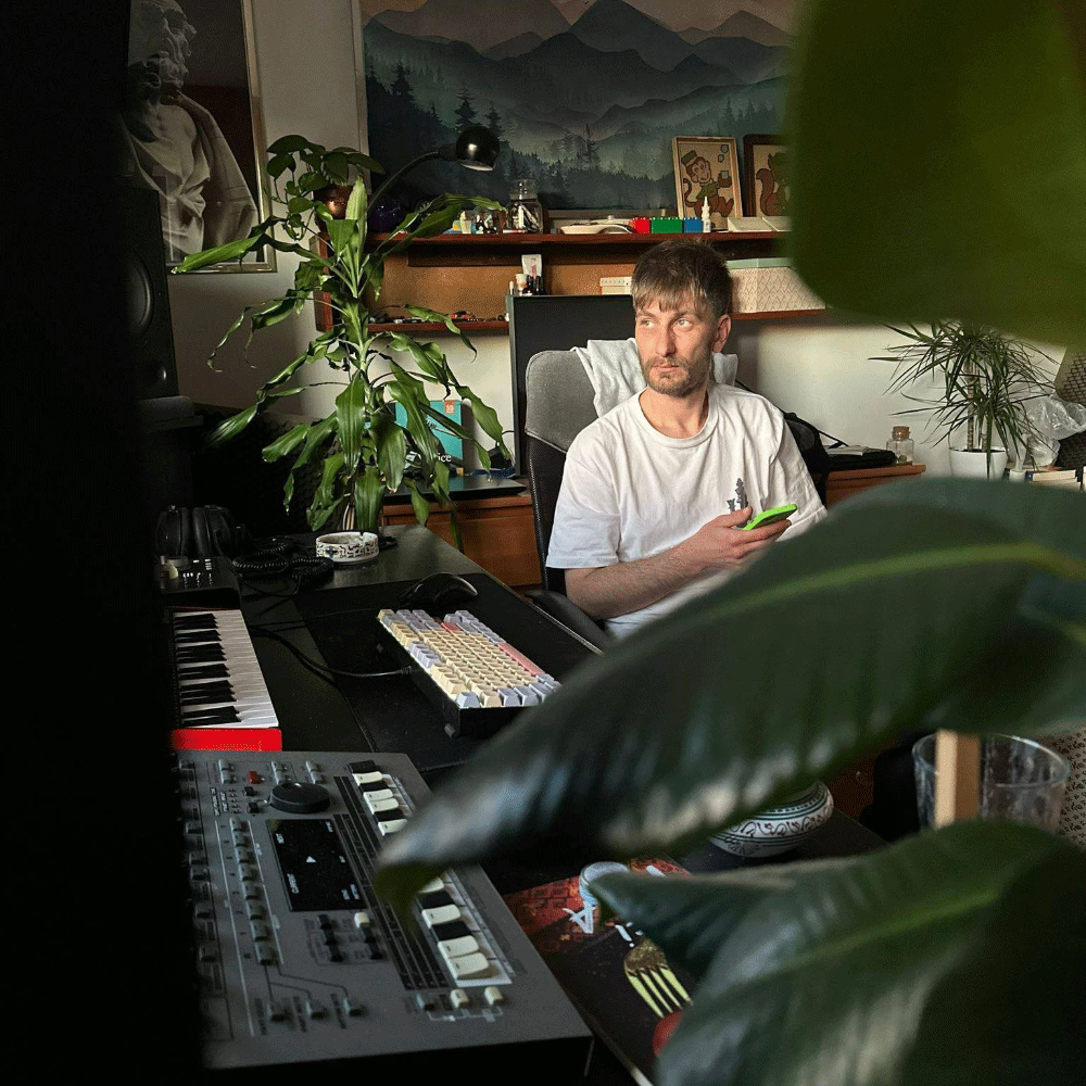 Man sitting in a music studio surrounded by instruments and plants, holding a green phone and looking to the side.