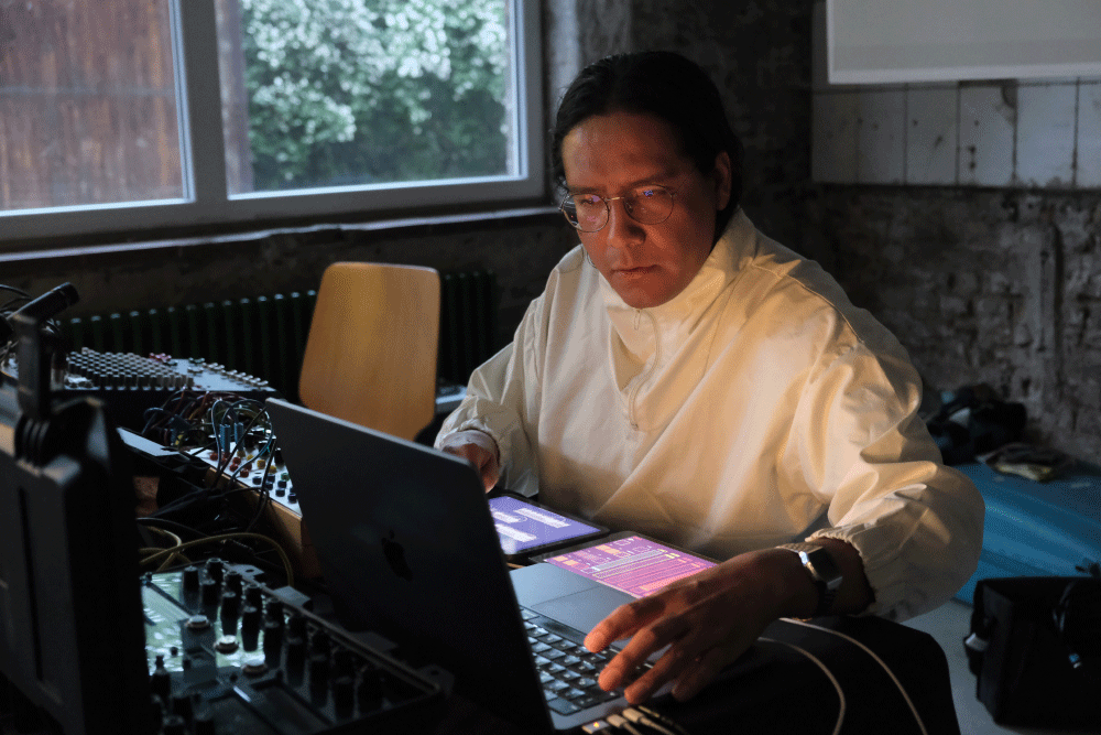 Person with glasses and a white jacket working on a laptop surrounded by audio equipment in a dimly lit room.