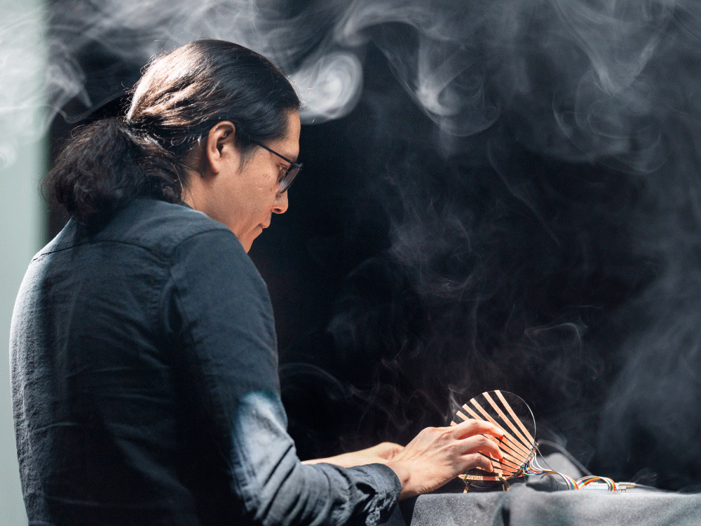 Person with long dark hair and glasses adjusting a curved electronic device on a table with smoke in the background.
