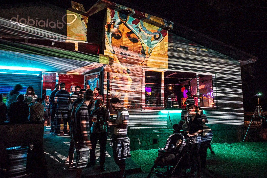 People gathered outside a brightly lit building at night with colorful projected artwork on the walls.