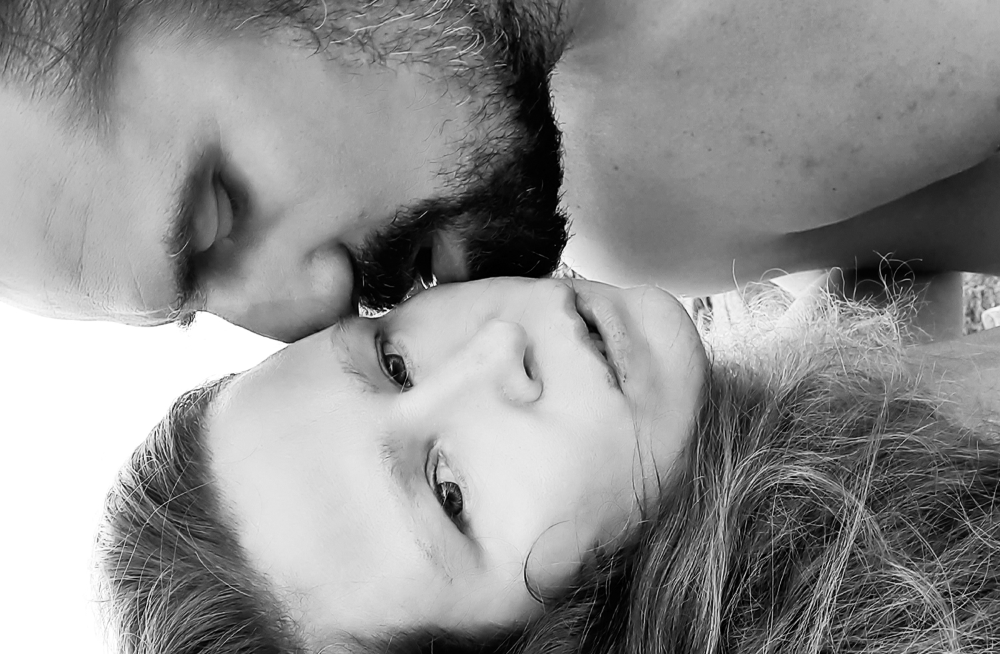 Black and white close-up of a man kissing a woman's forehead while she looks at the camera.