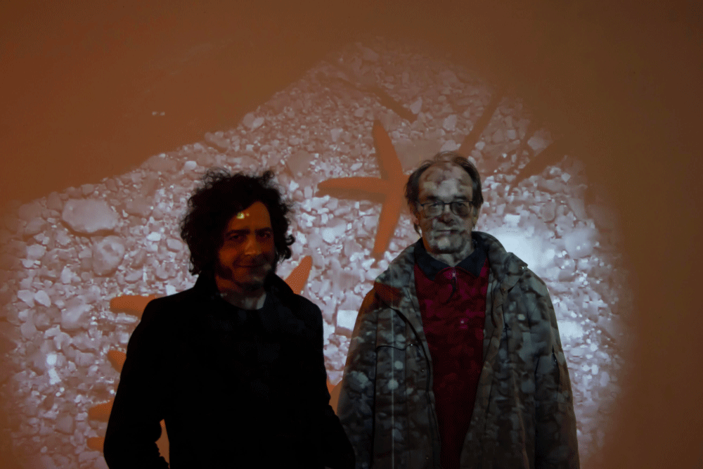Two men standing side by side with a projection of orange starfish and white sea stones overlayed on them and the background.