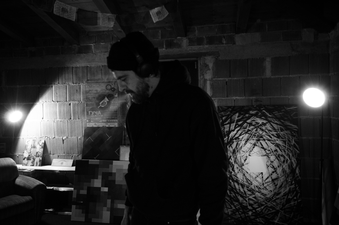 Black-and-white photo of a man wearing a beanie and headphones standing in a dimly lit room with pixelated and abstract artworks on the wall.