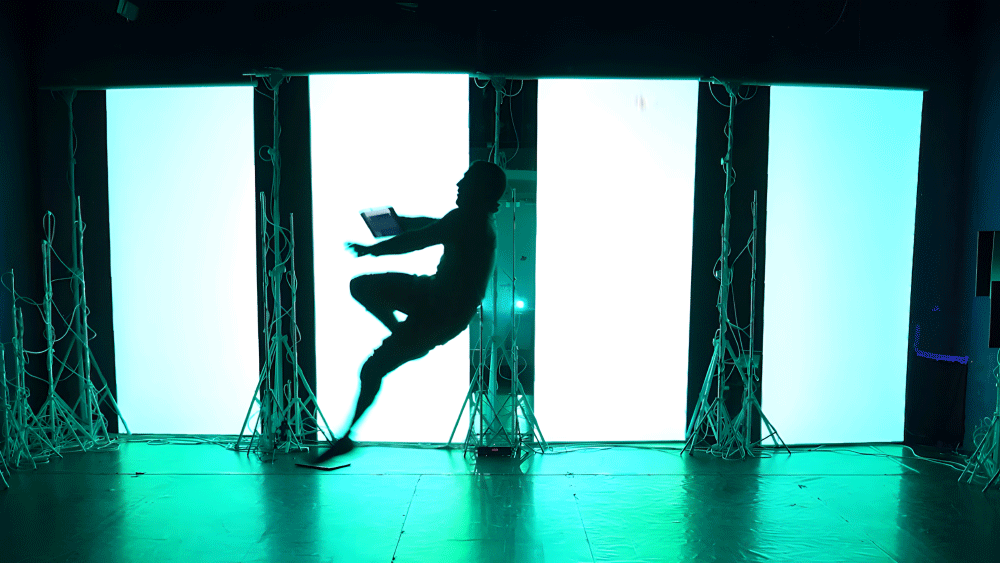 Silhouette of a person mid-air in front of bright, large vertical rectangular light panels with cables on the floor.
