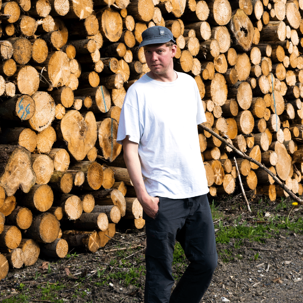 Man wearing a white t-shirt and black cap standing in front of a large stacked pile of cut logs outdoors.