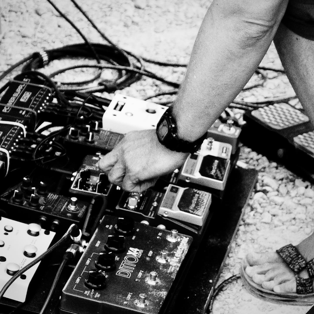 Person adjusting knobs on a pedalboard with various guitar effect pedals on a gravel surface, wearing a wristwatch and sandals.