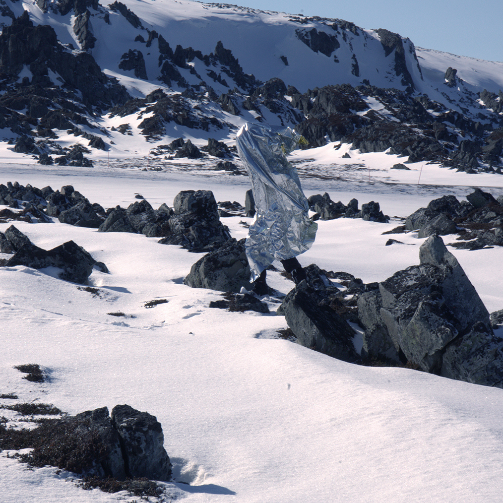 Person wrapped in a reflective emergency blanket walking on snowy rocky terrain with snow-covered mountains in the background.