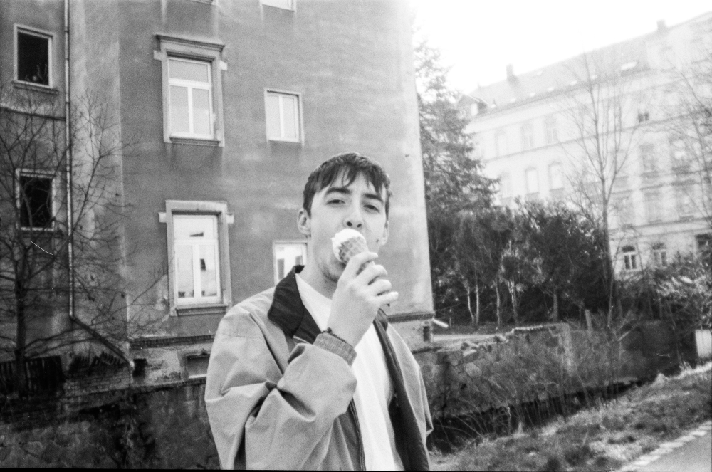 Young man wearing a jacket eating an ice cream cone standing outdoors near buildings and trees.