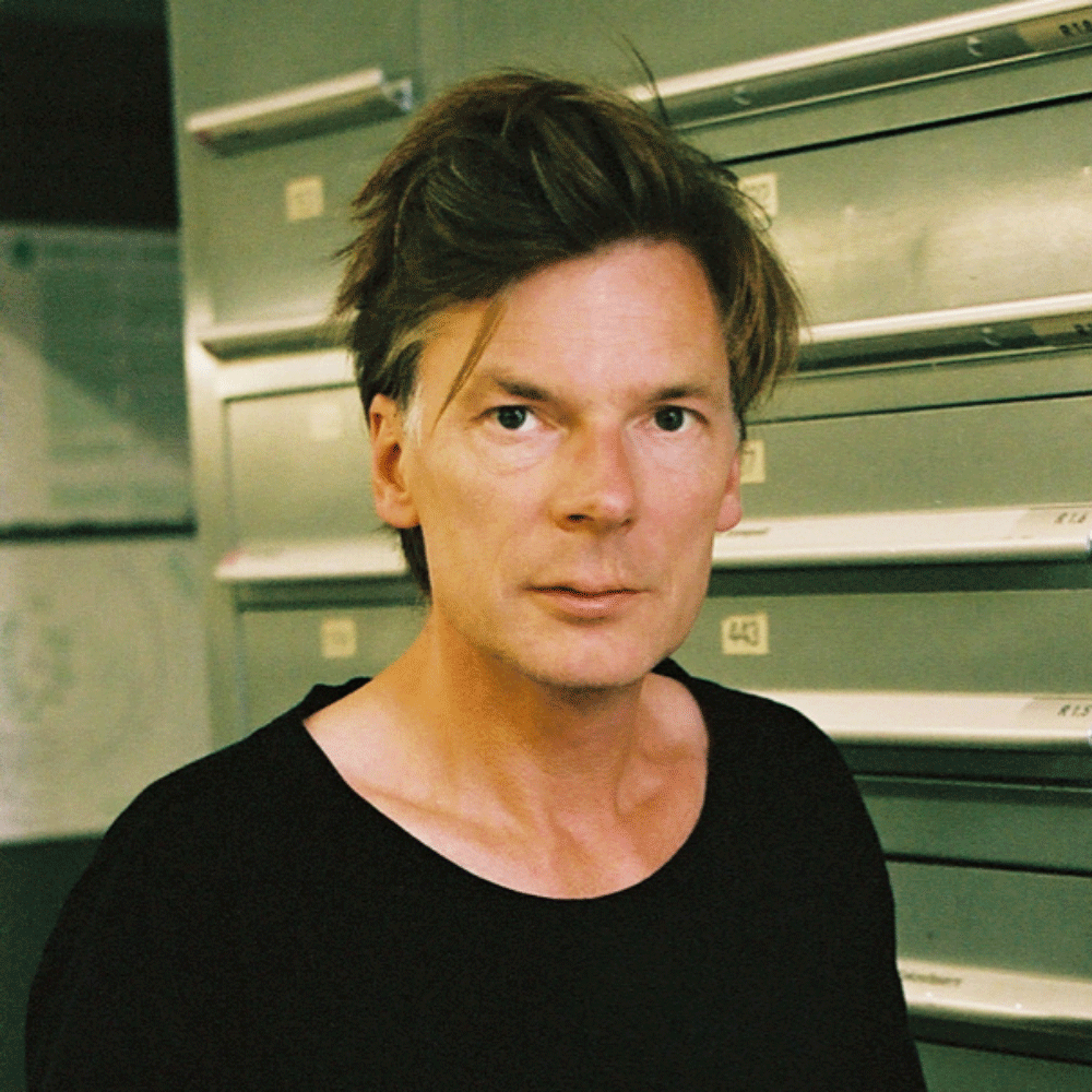 Man with tousled brown hair wearing a black shirt standing indoors in front of green lockers or mailboxes.