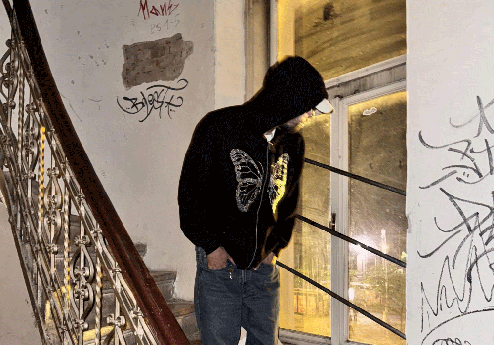 Person wearing a black hoodie with butterfly design and jeans standing inside near a graffiti-covered wall and ornate metal staircase railing.