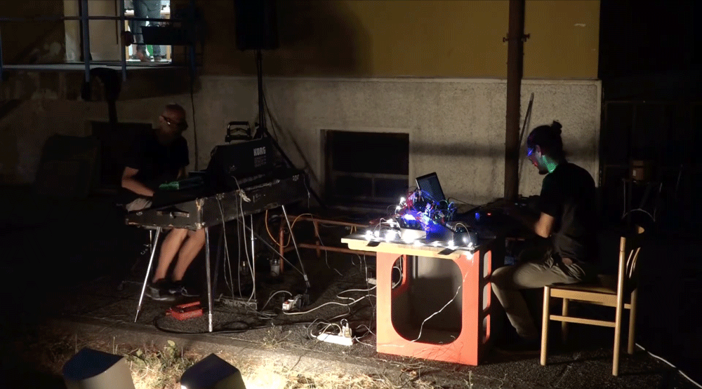 Two musicians perform outdoors at night, one playing a keyboard and the other working with electronic equipment on a table illuminated by string lights.