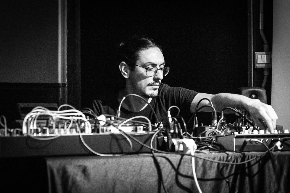 Man with glasses adjusting electronic music equipment with cables on a table in a dark room.