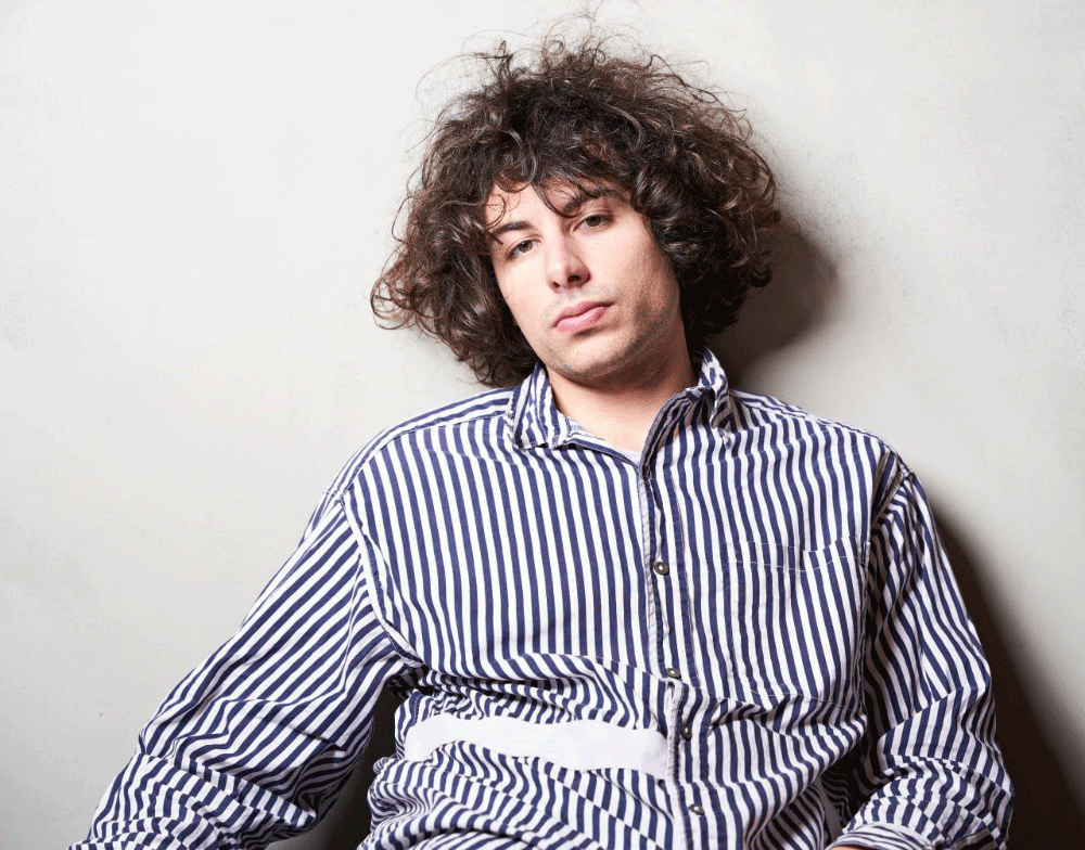 Young man with curly hair wearing a blue and white striped shirt, sitting against a light gray wall with a neutral expression.