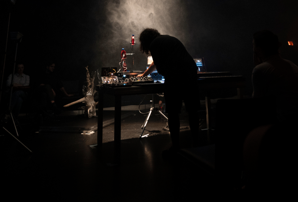Person adjusting electronic music equipment on a table with smoke and dim lighting in a dark room.