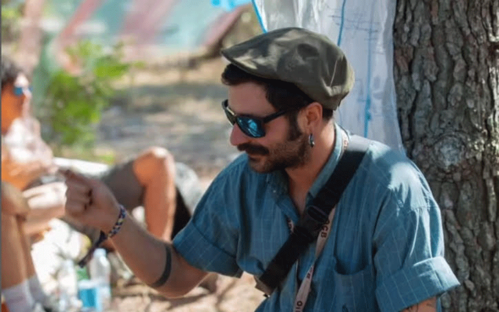 Man wearing a blue shirt, flat cap, and sunglasses sitting outdoors, smiling and extending his hand.