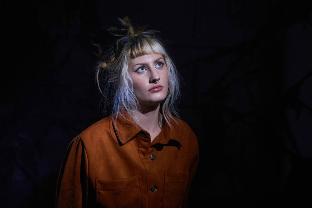 Young woman with blonde hair and bangs, wearing an orange jacket, looking upward against a dark background.