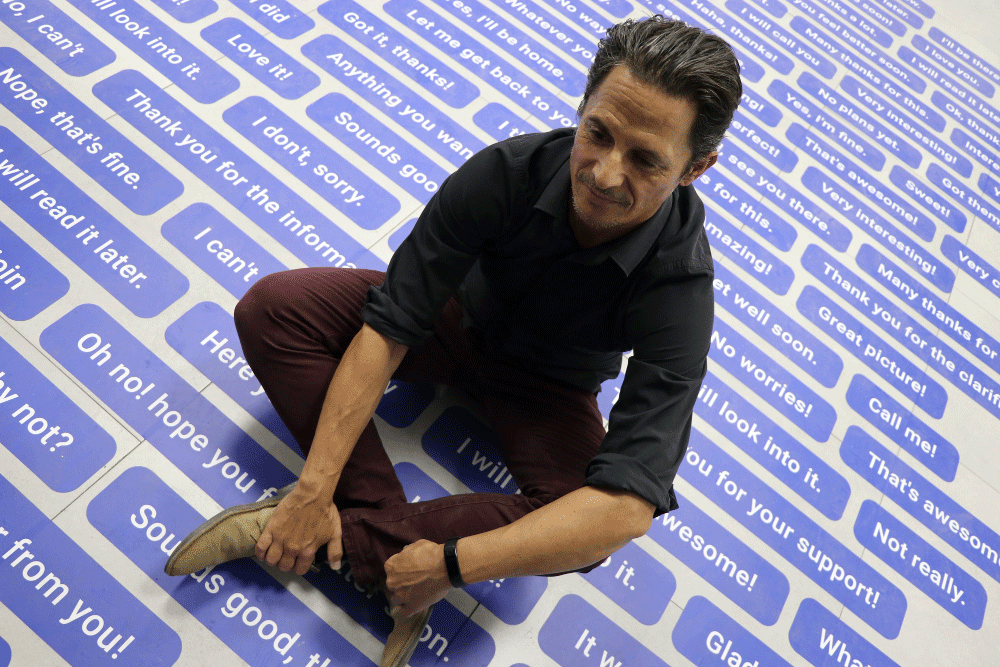 Man sitting cross-legged on a floor covered with blue and white speech bubble-style text boxes containing polite phrases.