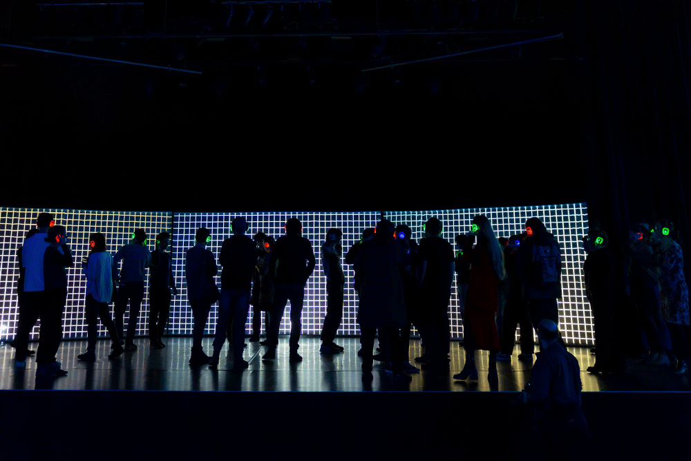 Group of people wearing colorful glowing headphones standing in front of a large illuminated grid backdrop in a dark room.