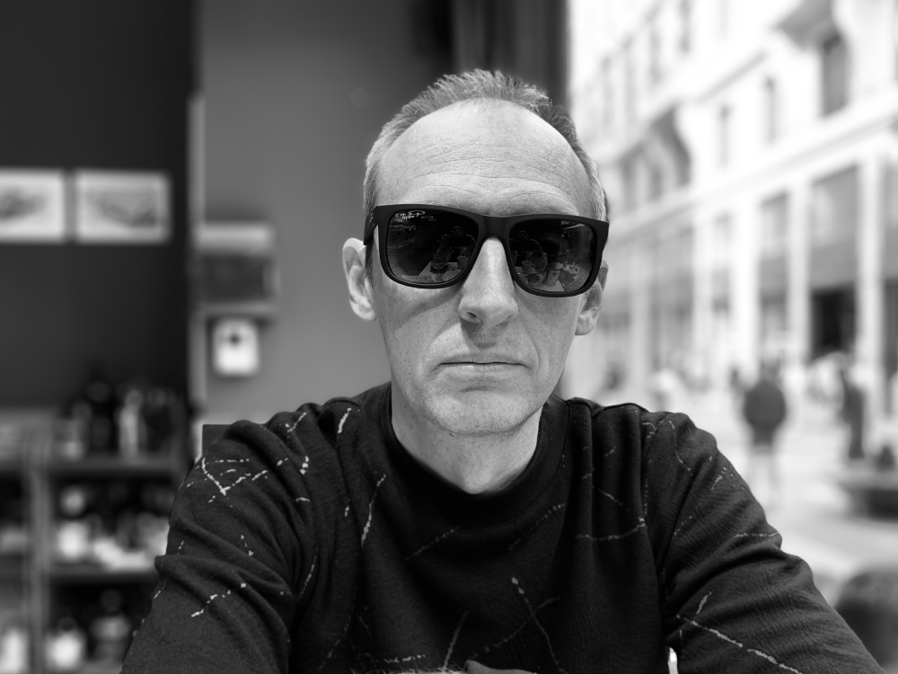 Black and white photo of a man wearing dark sunglasses and a patterned sweater, sitting indoors with a blurred urban street visible through the window behind him.