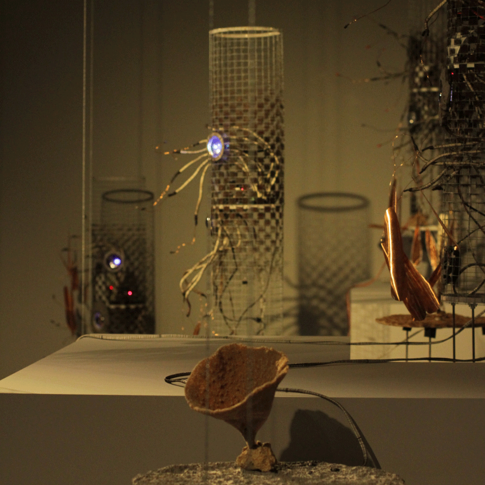 Dimly lit art installation featuring hanging cylindrical mesh structures with illuminated circular elements and wires, and a rough-textured, upside-down funnel-shaped object in the foreground.