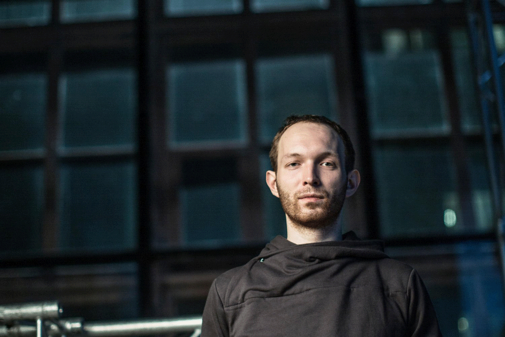 Portrait of a man with short brown hair and beard standing outdoors in front of a building with large windows.